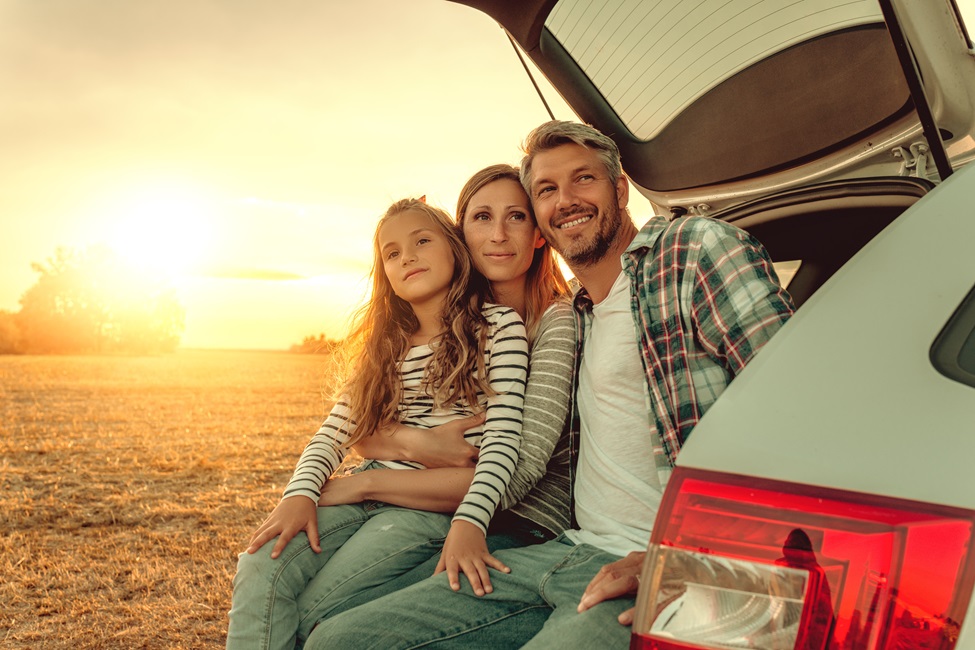 Eine Familie (Vater, Mutter, Tochter) sitzen im geöffneten Kofferraum ihres Autos und schaue in die Landschaft,