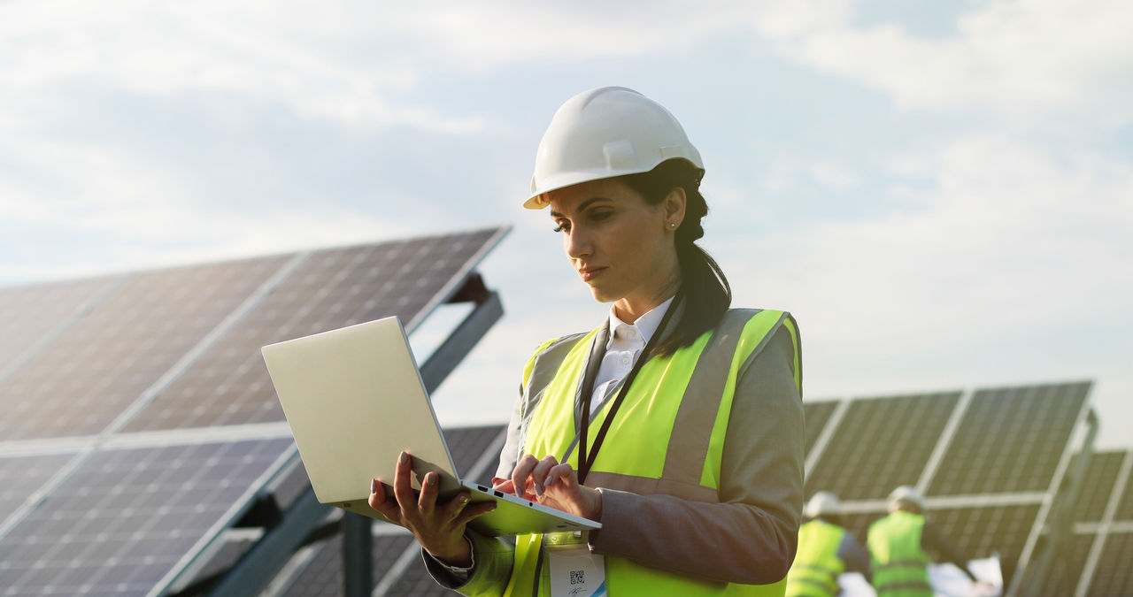 Porträt einer Elektrikerin in Schutzhelm und Arbeitskleidung, die mit einem Laptop Solarmodule prüft.