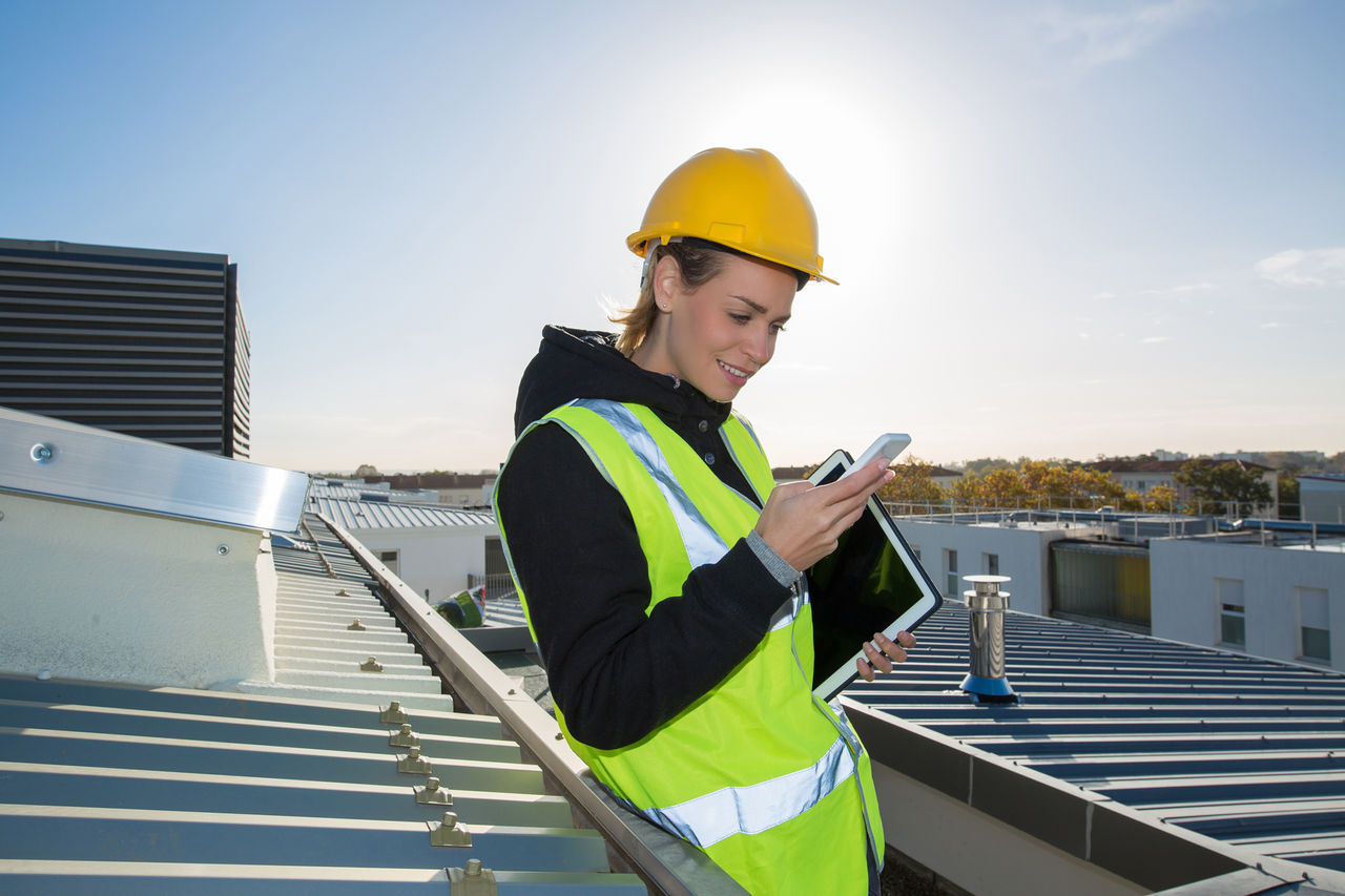 Eine Bauingenieurin auf dem Dach einer Baustelle mit Handy und Tablet.