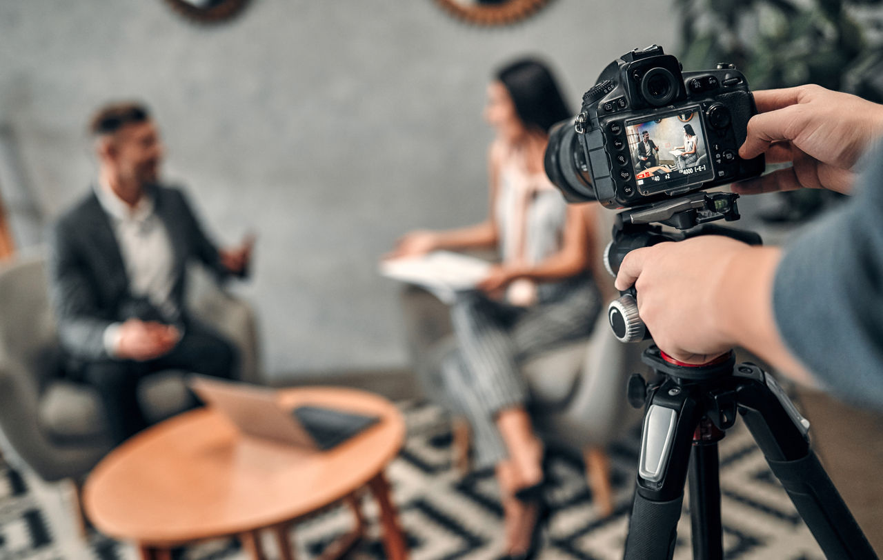 Auf dem Bild ist im Vordergrund eine Kamera zusehen, die ein Interview aufnimme, welches im Hintergrund zu sehen ist.
