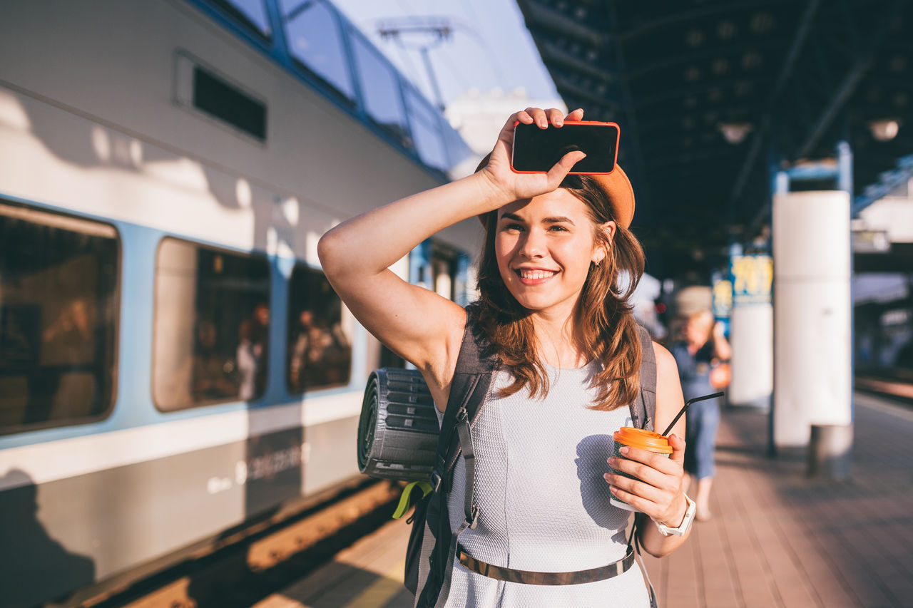 Eine junge Frau steht mit Rucksack, Handy und Kaffee in der Hand am Bahngleis.