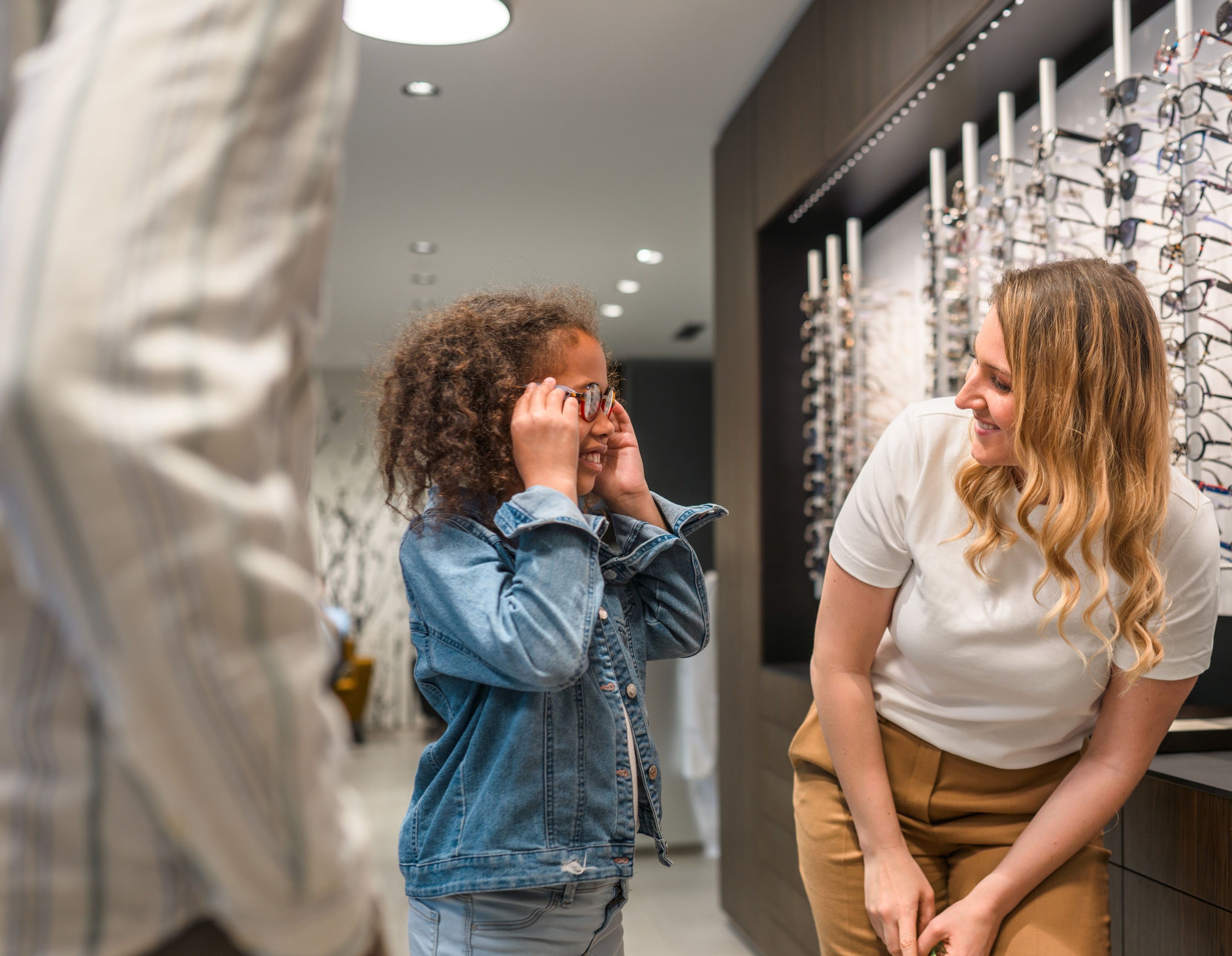 Schöne und modische Frau wählt Brillengestell in einem modernen Optikergeschäft. Verkäuferin hilft ihr, die richtige Entscheidung zu treffen.