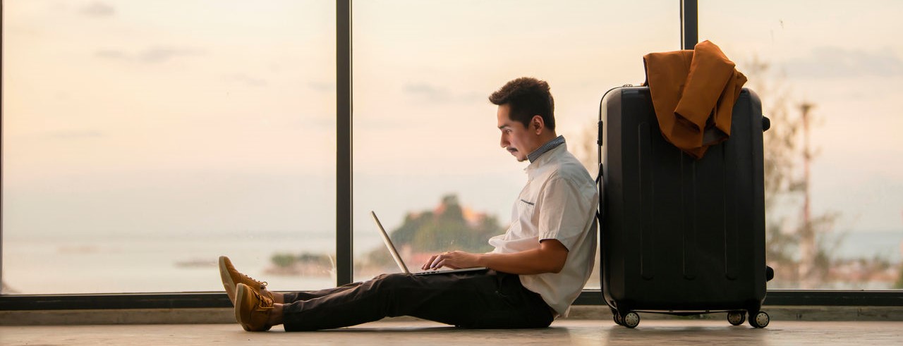 Ein junger Mann sitzt vor einer Aussicht am Laptop und lehnt sich an seinen Koffer.