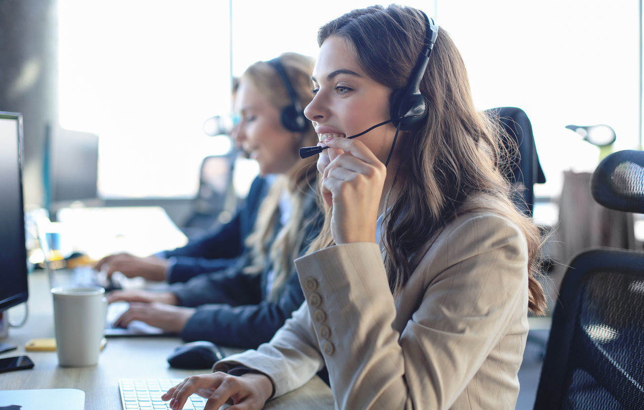 Kundenbetreuerin mit Headset bei der Arbeit. Im Hintergrund sitzen Ihre Kolleginnen und Kollegen.