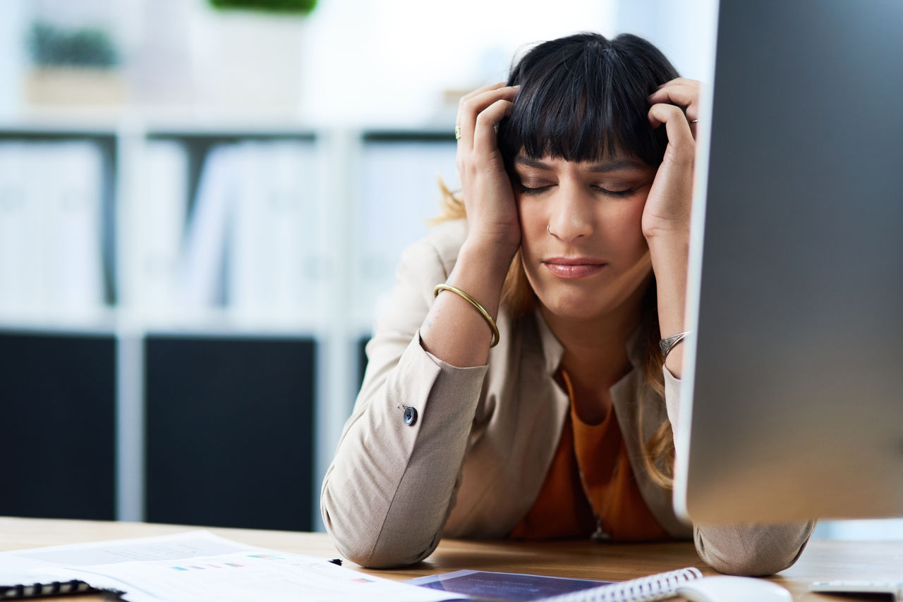 Eine Geschäftsfrau sitzt am Arbeitsplatz und greift sich erschöpft an die Schläfen.