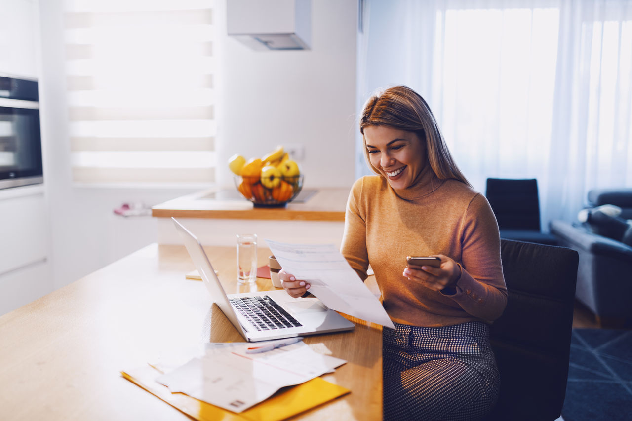 Eine Frau sitzt vor Ihrem Laptop mit Handy in der Hand an Ihrem Schreibtisch.