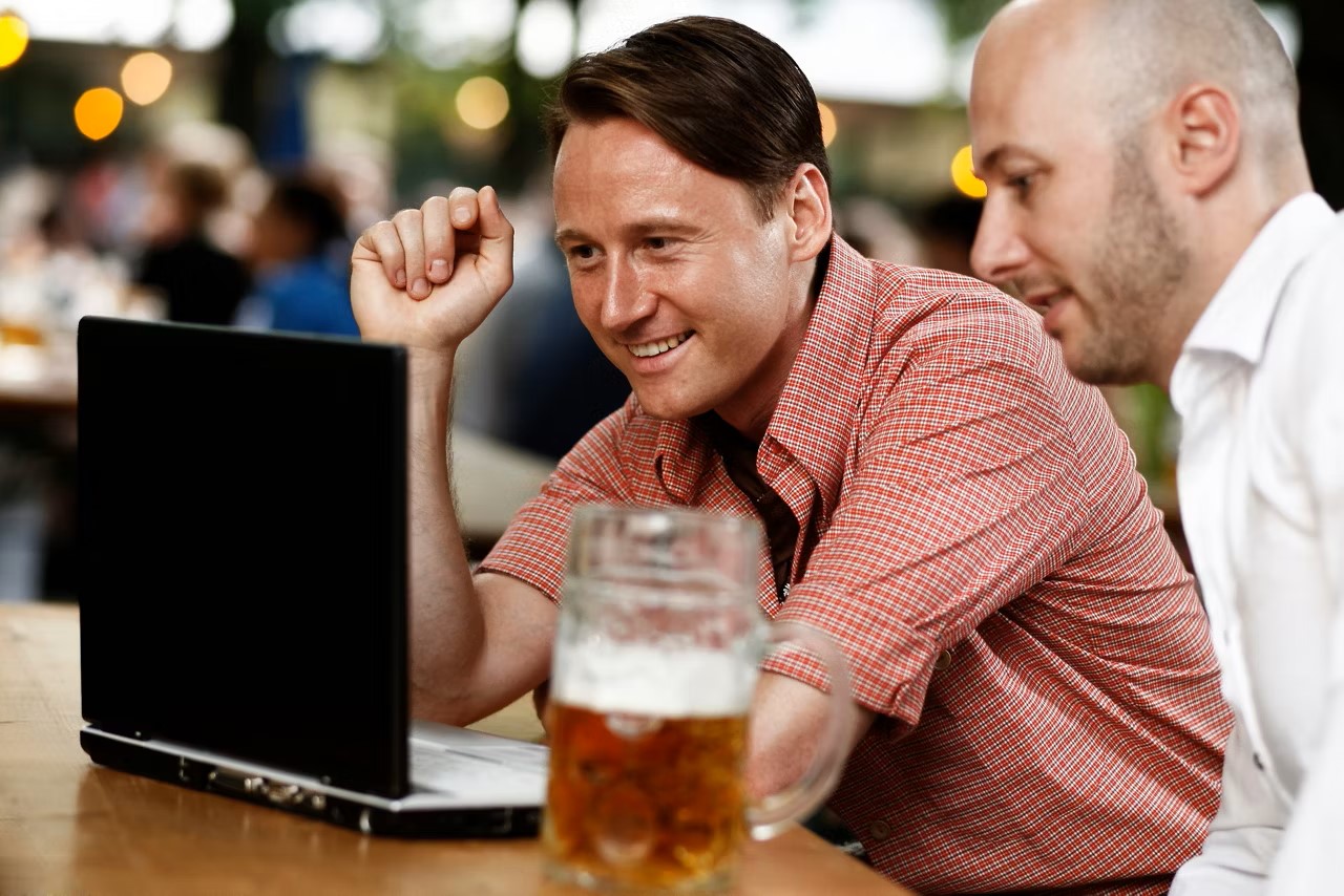 Zwei Männer schauen auf einen Laptop. Daneben steht eine Maa Bier.